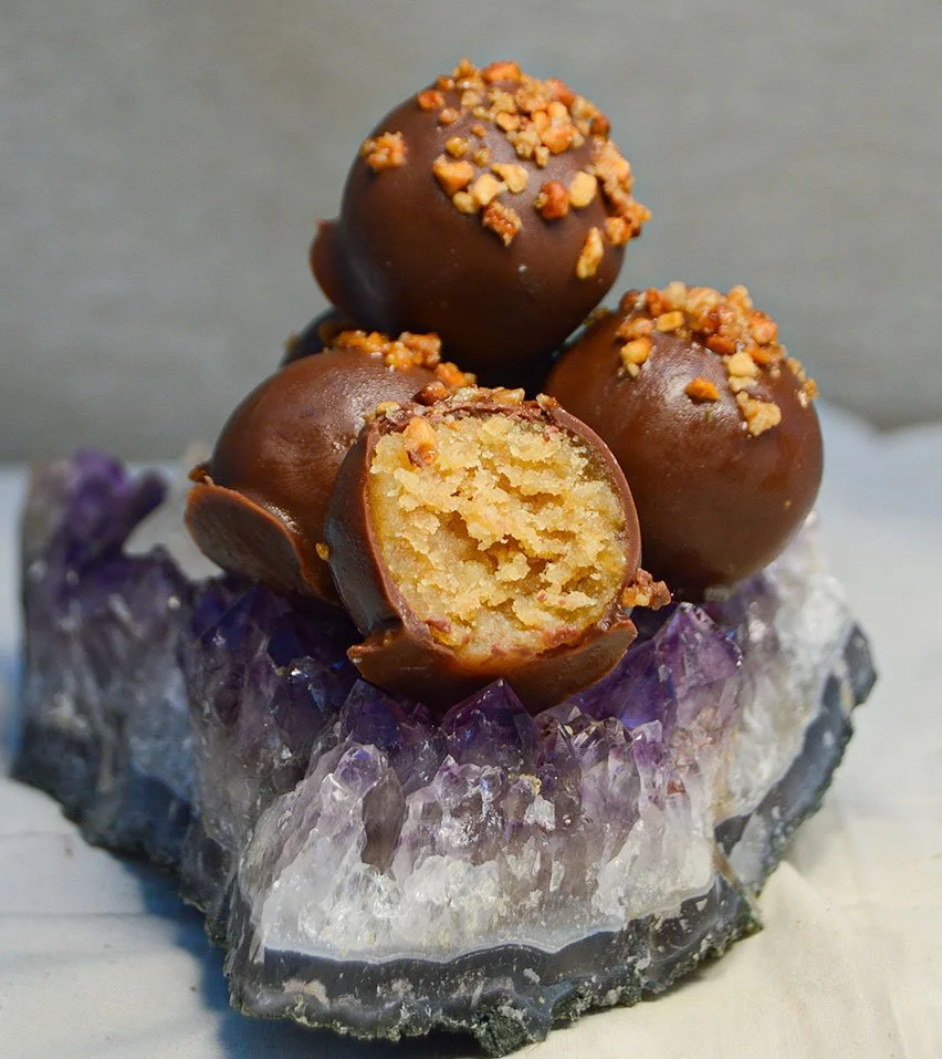 Imagen de los delis de crema de cacahuete, que son bolitas de un solo bocado de masa de bizcocho con distintos sabores, cubiertos con chocolate y decorados. Uno de ellos está partido por la mitad y se puede ver su textura interior.