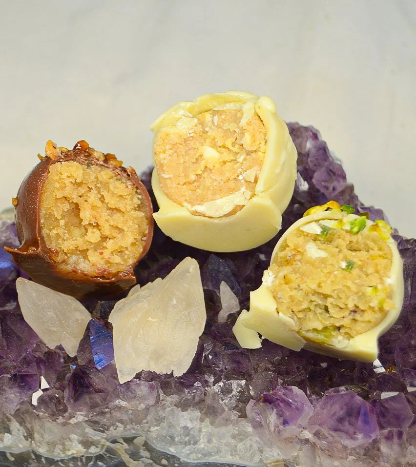 Imagen de los delis de varios sabores, que son bolitas de un solo bocado de masa de bizcocho con distintos sabores, cubiertos con chocolate y decorados. 3 de ellos están partidos por la mitad y se puede ver su textura interior.