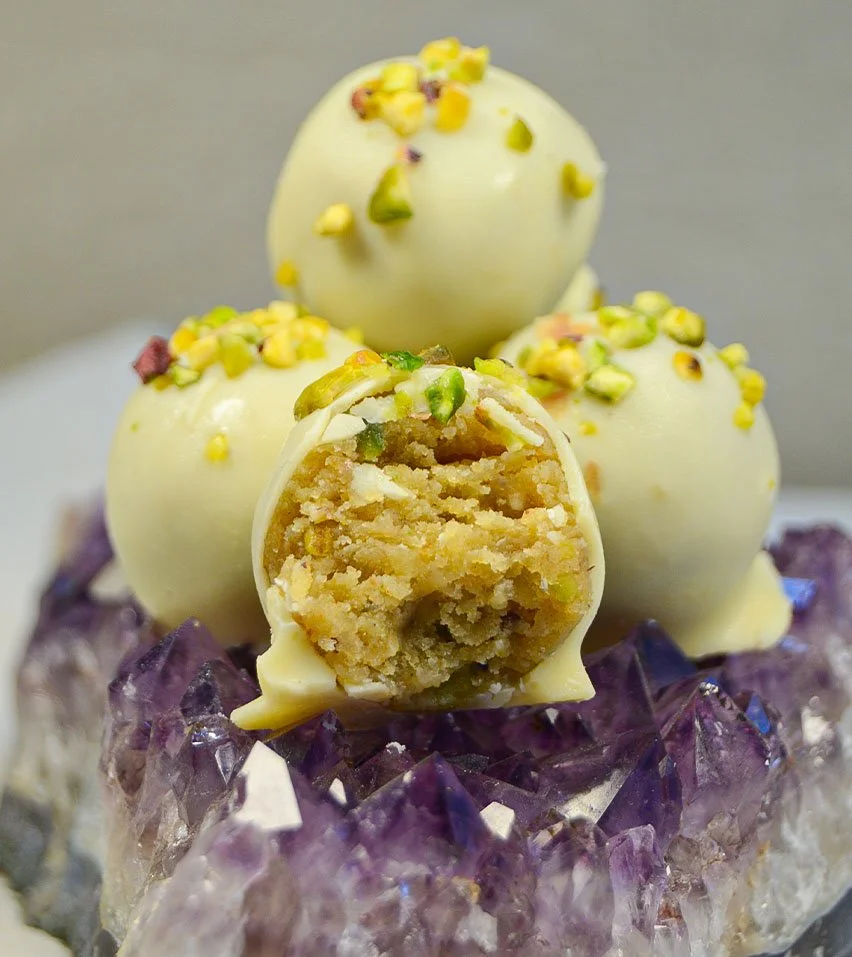 Imagen de los delis de pistacho, que son bolitas de un solo bocado de masa de bizcocho con distintos sabores, cubiertos con chocolate y decorados. Uno de ellos está partido por la mitad y se puede ver su textura interior.