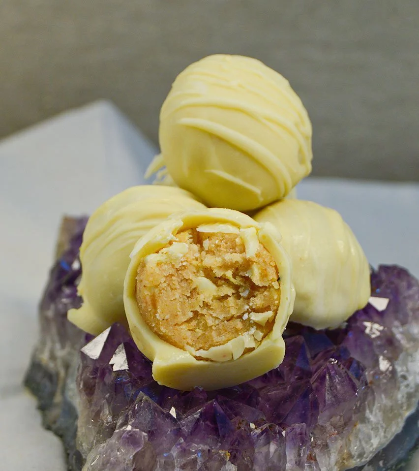 Imagen de los delis de chocolate blanco con vainilla, que son bolitas de un solo bocado de masa de bizcocho con distintos sabores, cubiertos con chocolate y decorados. Uno de ellos está partido por la mitad y se puede ver su textura interior.